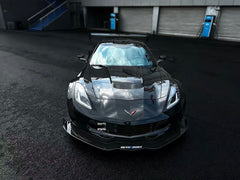 Black Chevrolet Corvette race car parked on a wet industrial track surface, featuring an aggressive wide-body kit, large rear wing, carbon fiber aerodynamic components, and lightweight performance wheels with red brake calipers. The moody urban backdrop and reflective pavement emphasize the car’s high-performance motorsport styling.