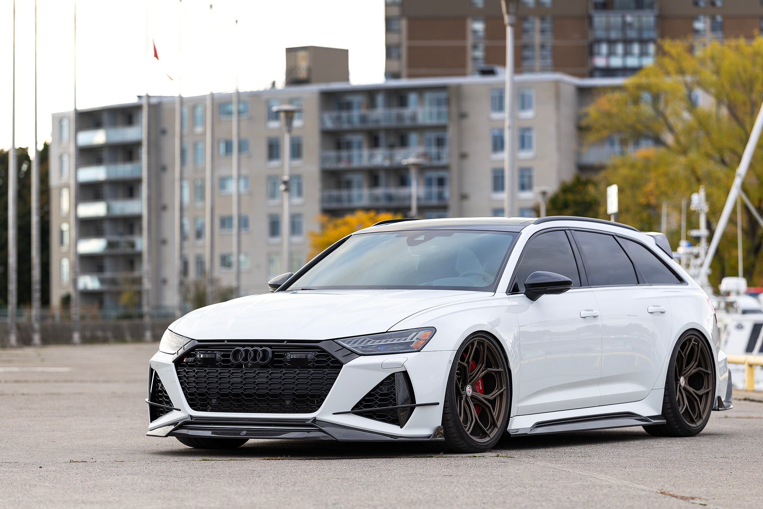 White Audi RS6 Avant featuring a REVOZPORT carbon fiber body kit with front lip, side skirts, rear diffuser, and lowered stance parked in a modern urban waterfront setting.