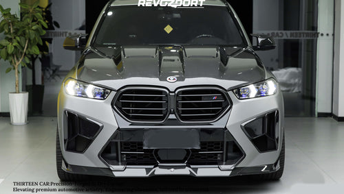 Front view of a white BMW X5M featuring a REVOZPORT carbon fiber hood, aggressive front bumper, enlarged air intakes, and carbon fiber front lip displayed inside a modern showroom.