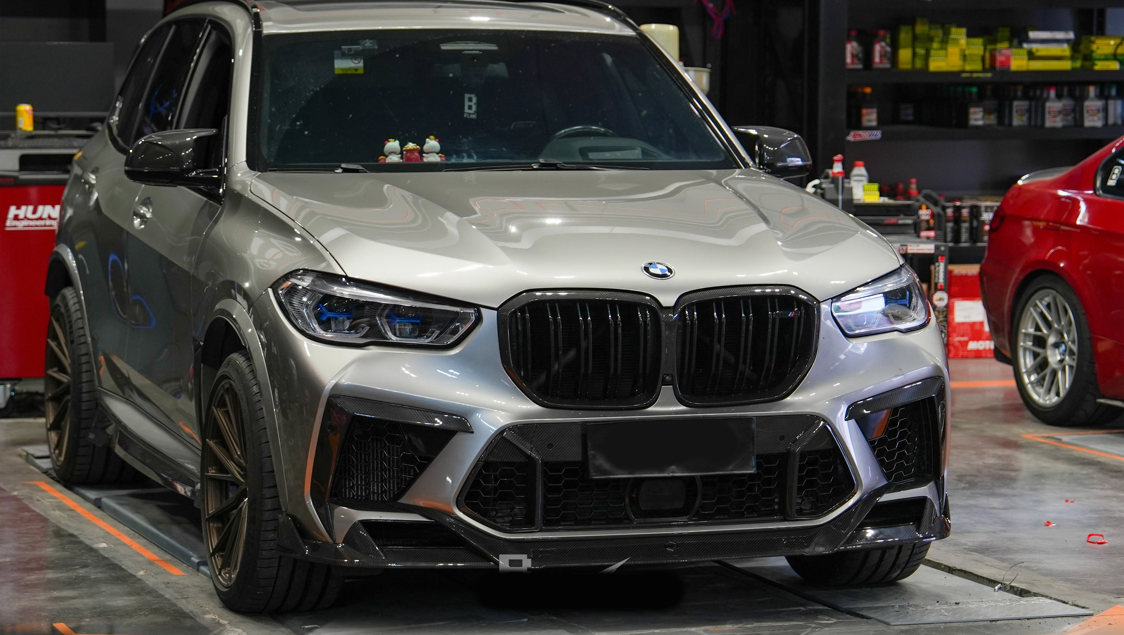 Gray BMW X5M featuring a REVOZPORT carbon fiber body kit with aggressive front lip, enlarged air intakes, side skirts, and sporty aero styling displayed inside a performance workshop.