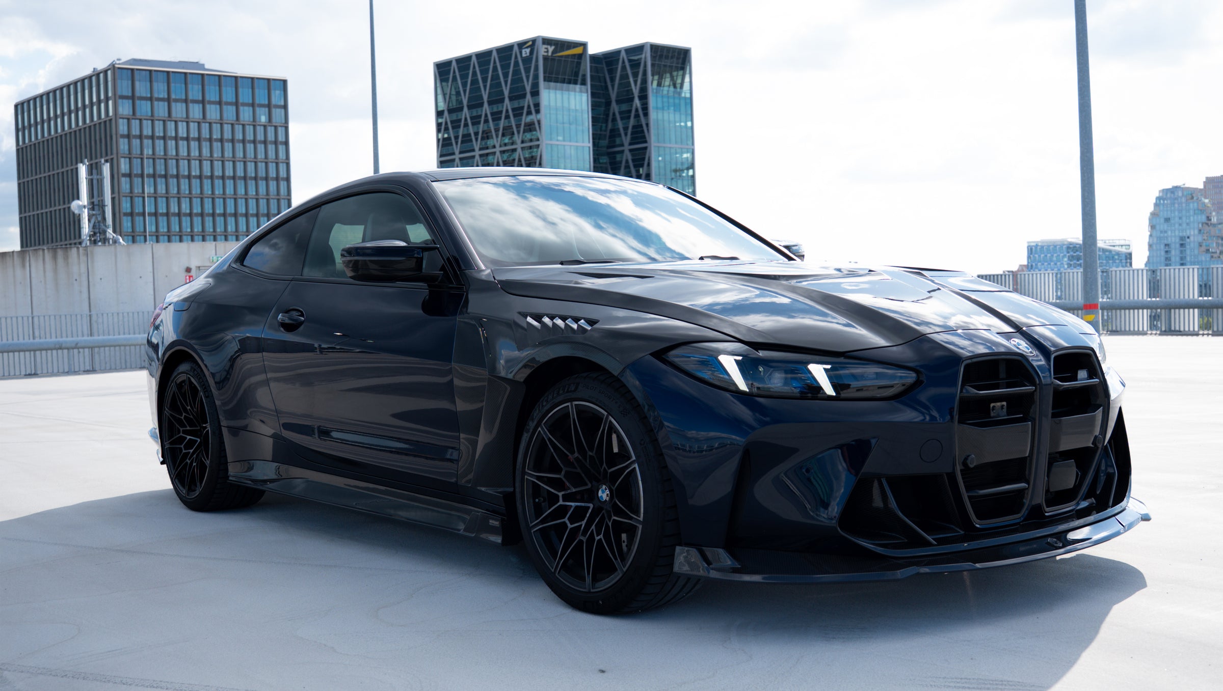 Black BMW G82 M4 featuring a REVOZPORT carbon fiber body kit with front lip, side skirts, vented fenders, and aggressive aero styling parked on a rooftop with city buildings in the background.