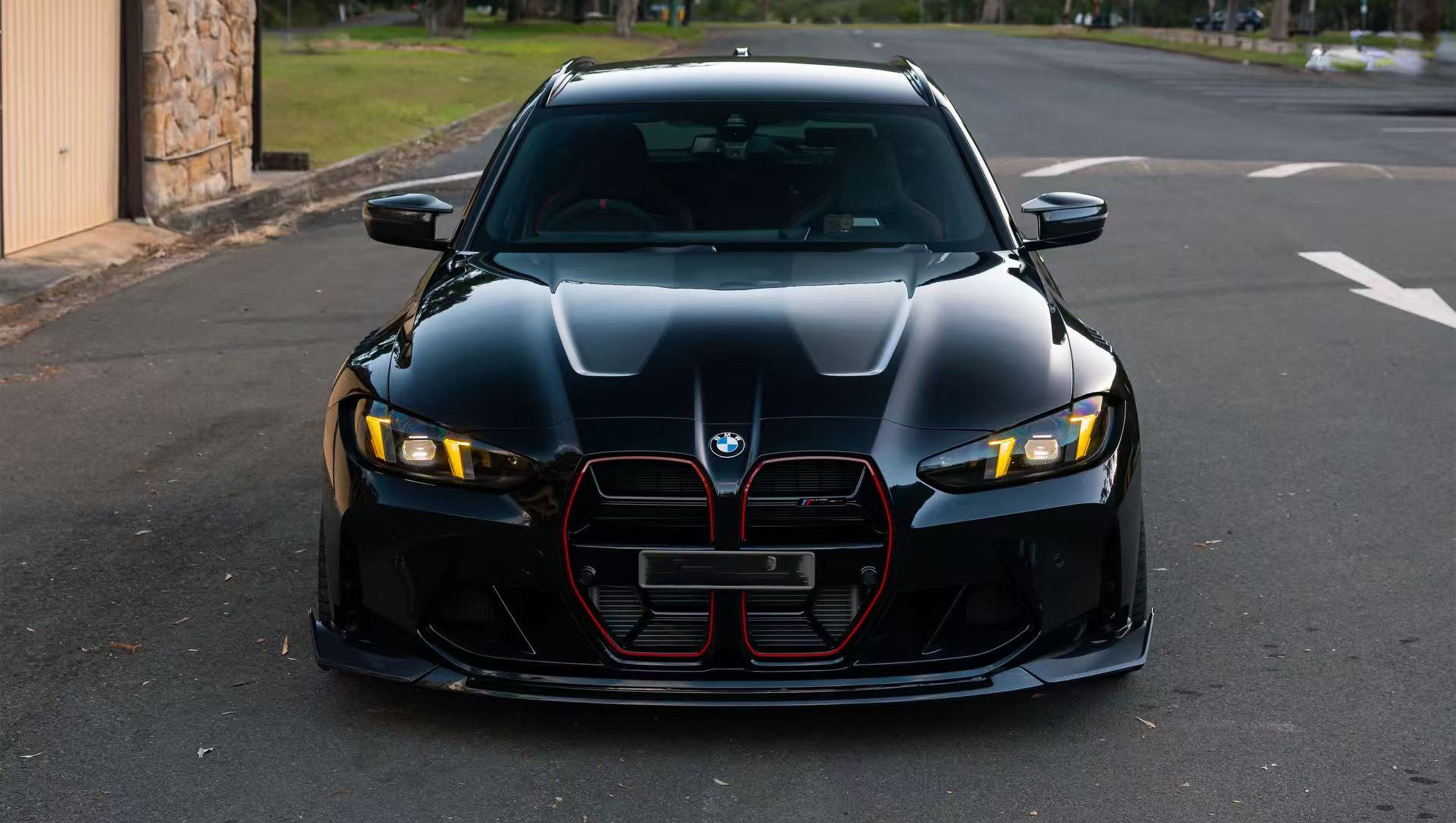 Black BMW G81 M3 Touring featuring a REVOZPORT carbon fiber body kit with vented fenders, side skirts, bronze wheels, and red performance brakes parked beside a stone wall.
