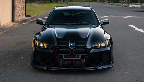 Black BMW G81 M3 Touring featuring a REVOZPORT carbon fiber body kit with vented fenders, side skirts, bronze wheels, and red performance brakes parked beside a stone wall.