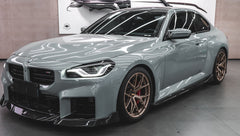 Gray BMW G87 M2 featuring a REVOZPORT carbon fiber front lip, aggressive front aero styling, bronze wheels, and red performance brakes displayed in a modern indoor garage.