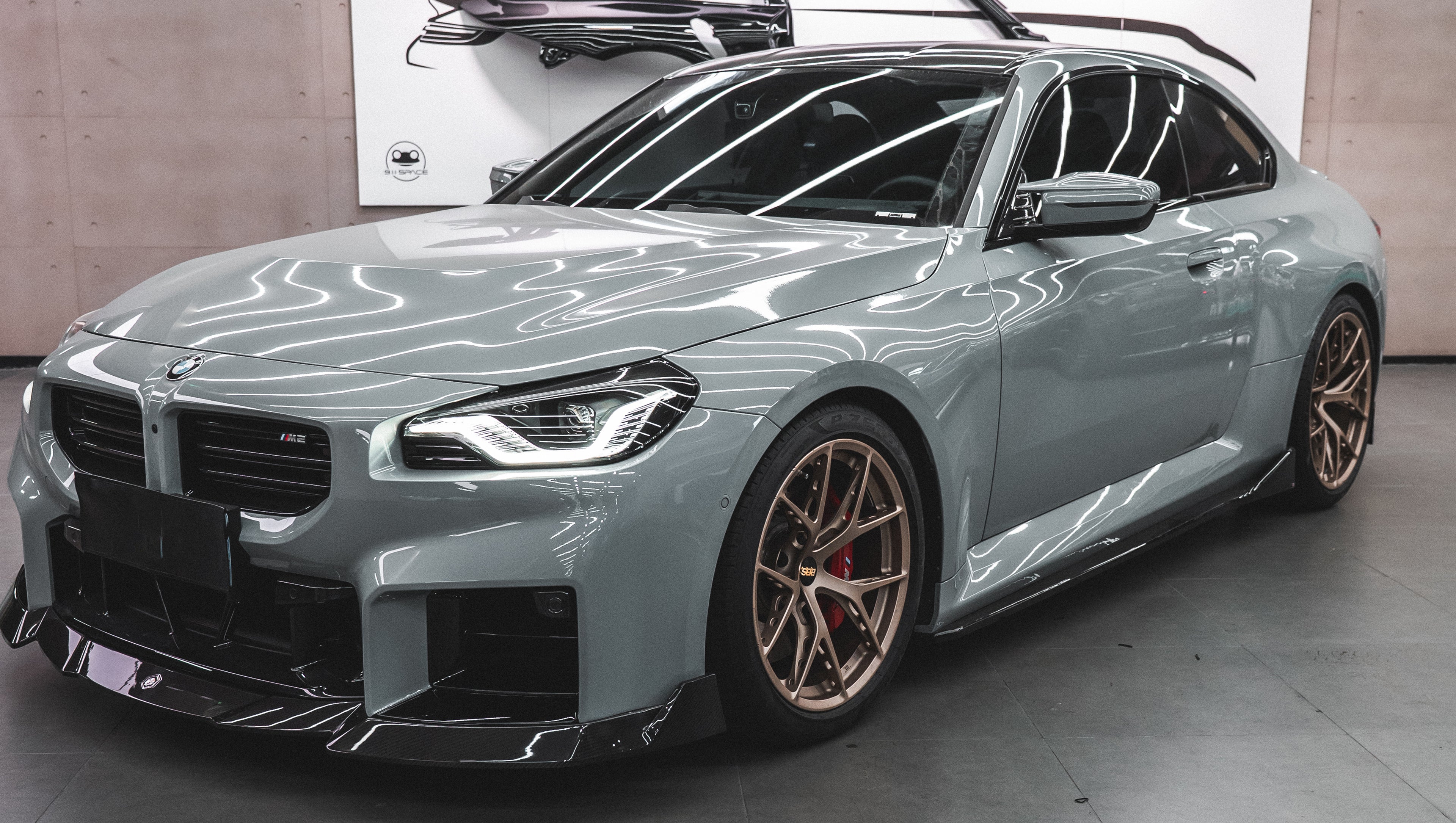 Gray BMW G87 M2 featuring a REVOZPORT carbon fiber front lip, aggressive front aero styling, bronze wheels, and red performance brakes displayed in a modern indoor garage.