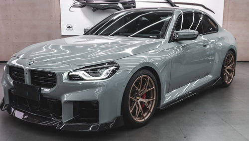 Gray BMW G87 M2 featuring a REVOZPORT carbon fiber front lip, aggressive front aero styling, bronze wheels, and red performance brakes displayed in a modern indoor garage.