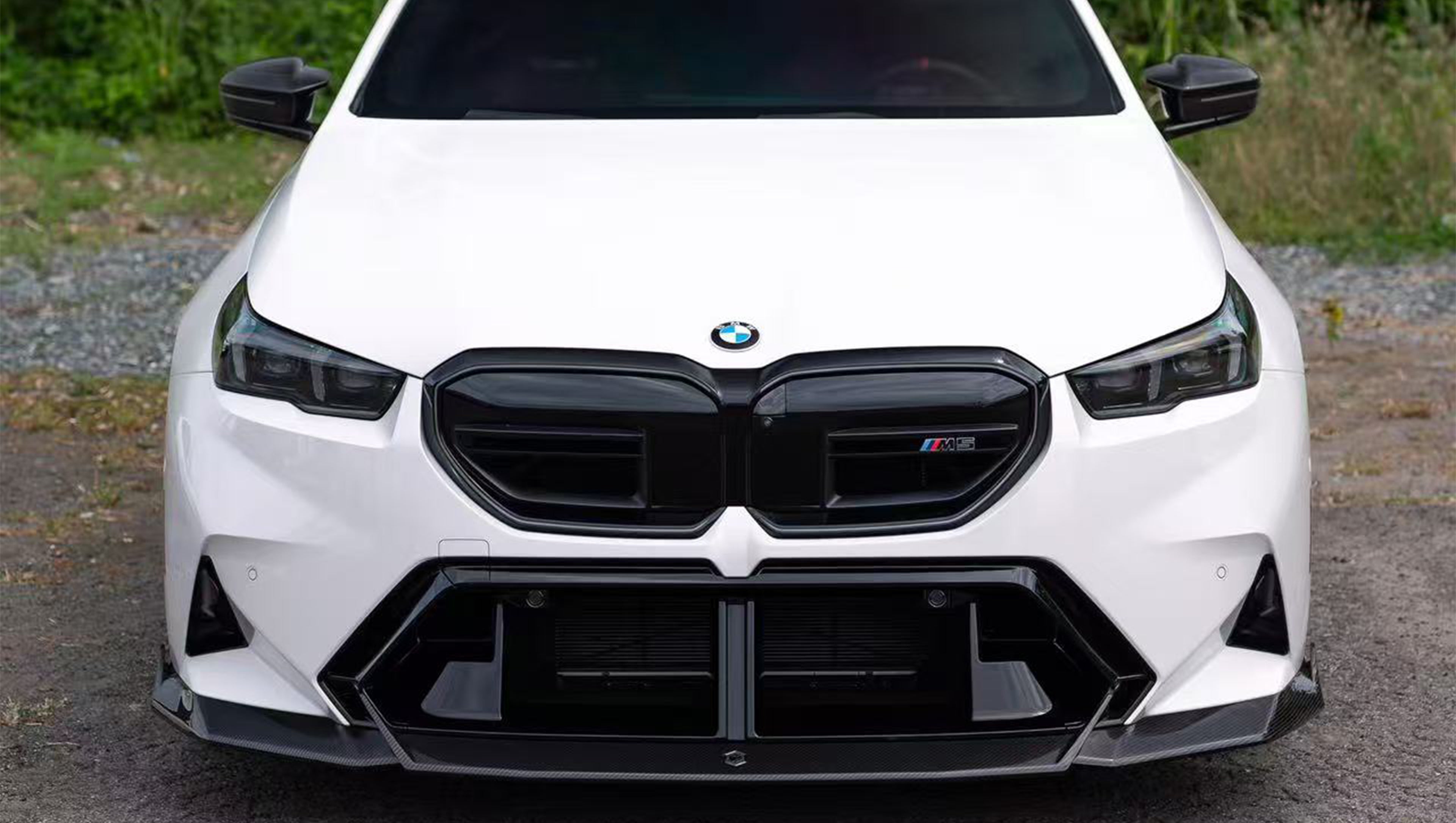 White BMW G90 M5 featuring a REVOZPORT carbon fiber front lip, side skirts, and aggressive front aero styling parked in a natural outdoor setting.
