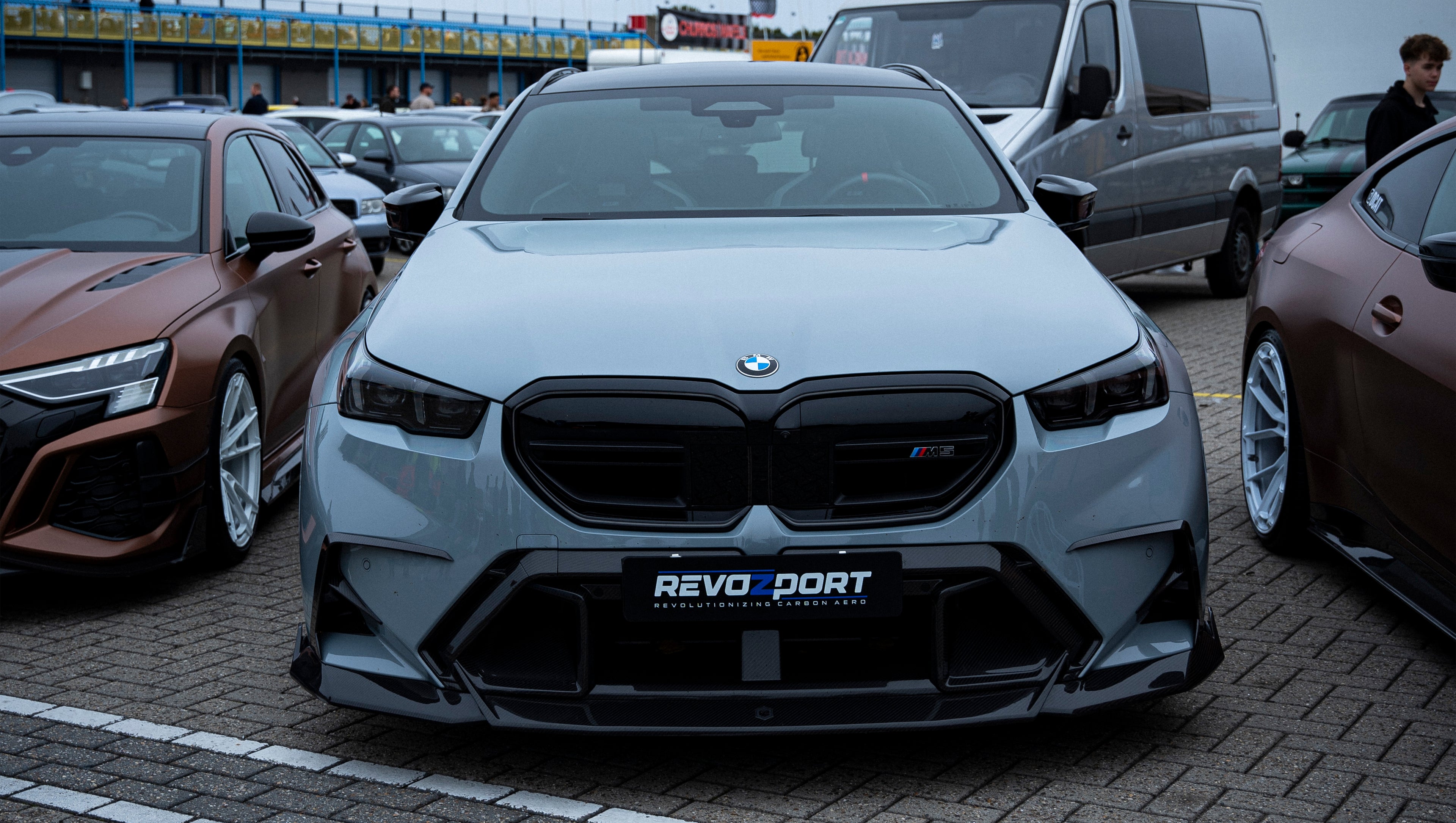 Gray BMW G90 M5 Touring featuring a REVOZPORT carbon fiber front lip, aggressive front aero styling, and black grille displayed at a car event.
