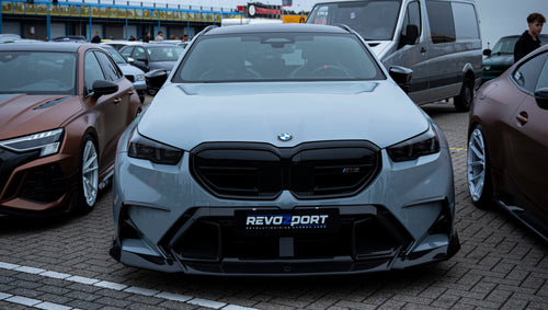 Gray BMW G90 M5 Touring featuring a REVOZPORT carbon fiber front lip, aggressive front aero styling, and black grille displayed at a car event.
