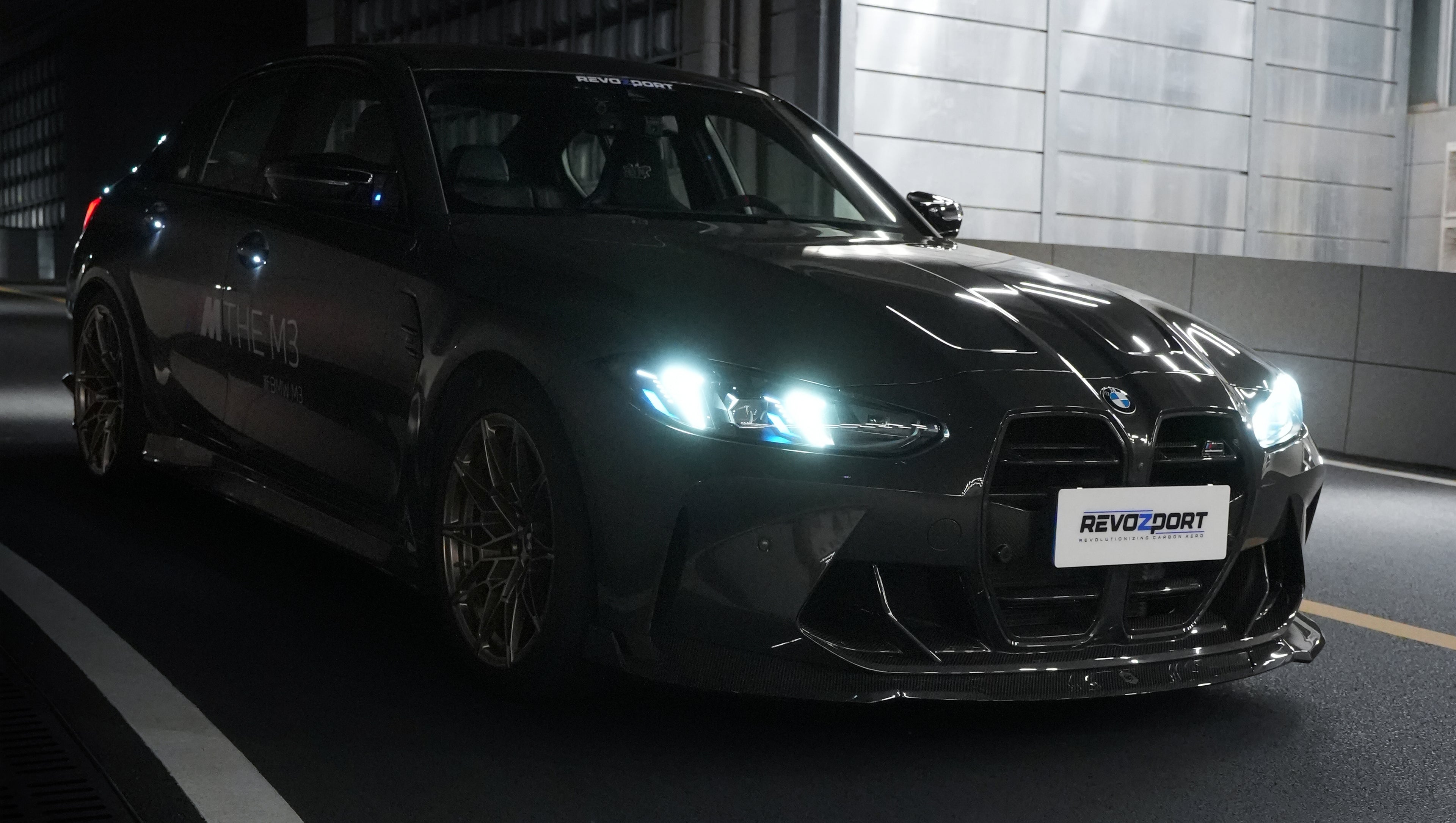 Gray BMW G80 M3 featuring a REVOZPORT carbon fiber front lip, side skirts, bronze wheels, and M The M3 side graphics driving through a tunnel at night.