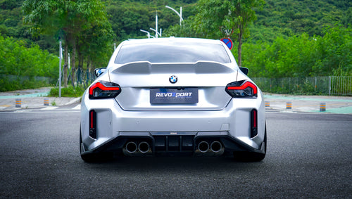 Close-up of a silver BMW G87 M2 featuring a REVOZPORT carbon fiber front lip, aggressive front aero styling, side skirt details, and white multi-spoke wheels parked in a modern urban setting.