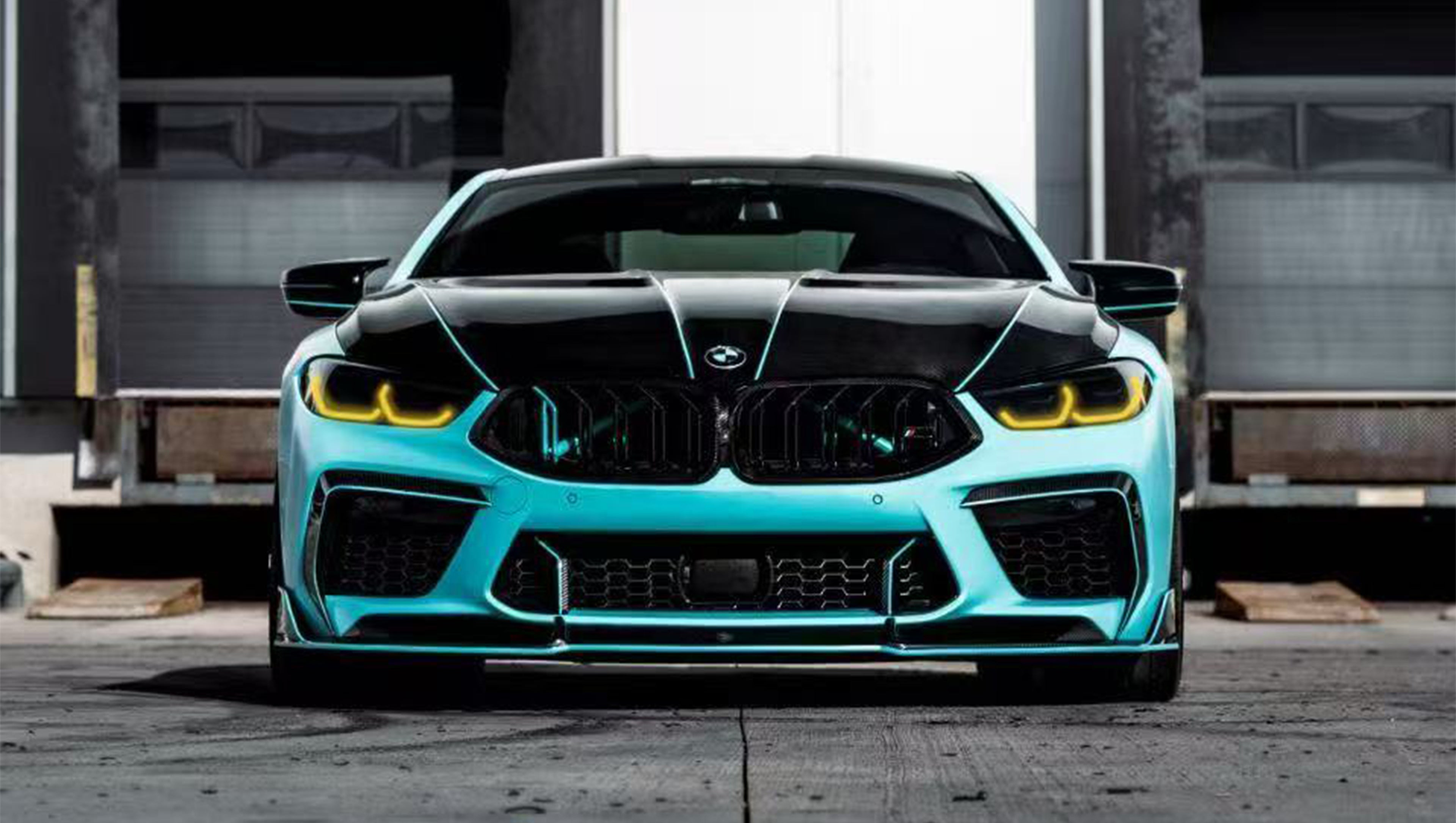 Turquoise BMW F92 M8 featuring a REVOZPORT carbon fiber body kit with front lip, side skirts, black hood accents, side vents, and silver forged wheels parked outside an industrial loading dock.