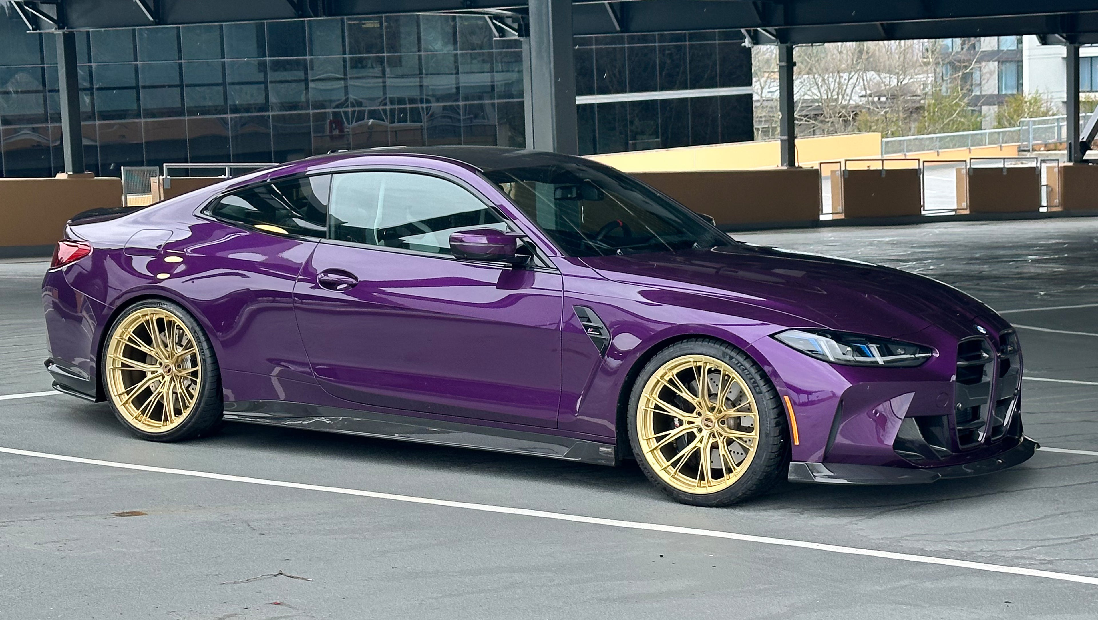 Purple BMW G82 M4 featuring a REVOZPORT carbon fiber body kit with front lip, side skirts, vented fenders, and gold wheels parked outside a modern industrial building.
