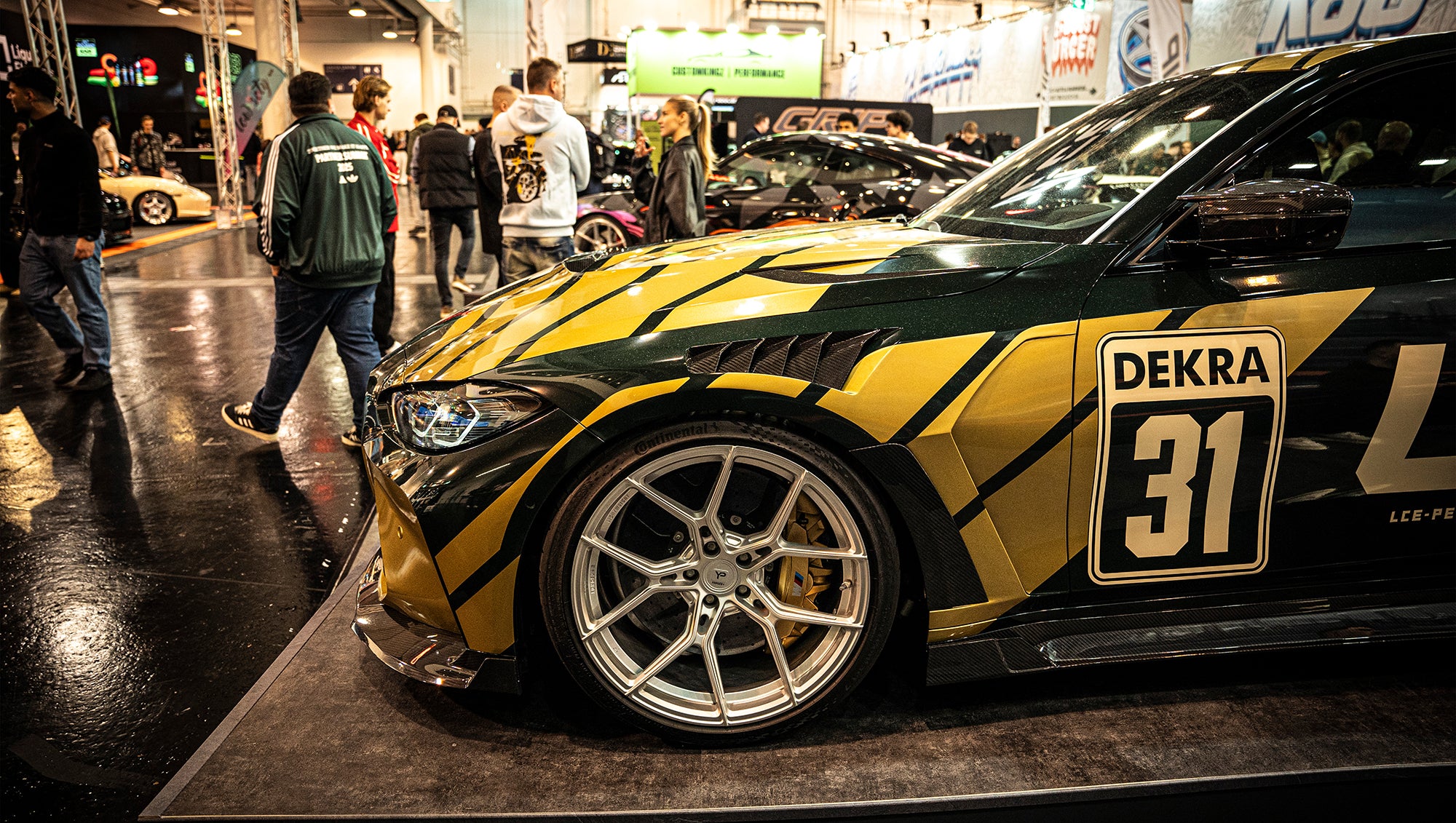BMW G80 M3 race build with REVOZPORT carbon fiber aero kit, gold wheels, and black-yellow motorsport livery displayed at an indoor automotive exhibition.