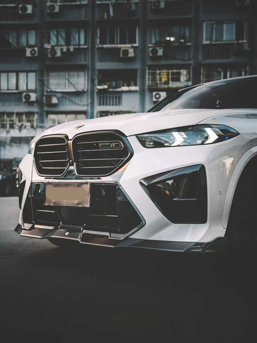 Front view of a white BMW X5M featuring a REVOZPORT carbon fiber front lip and aggressive front bumper aero styling parked on an urban street at night.