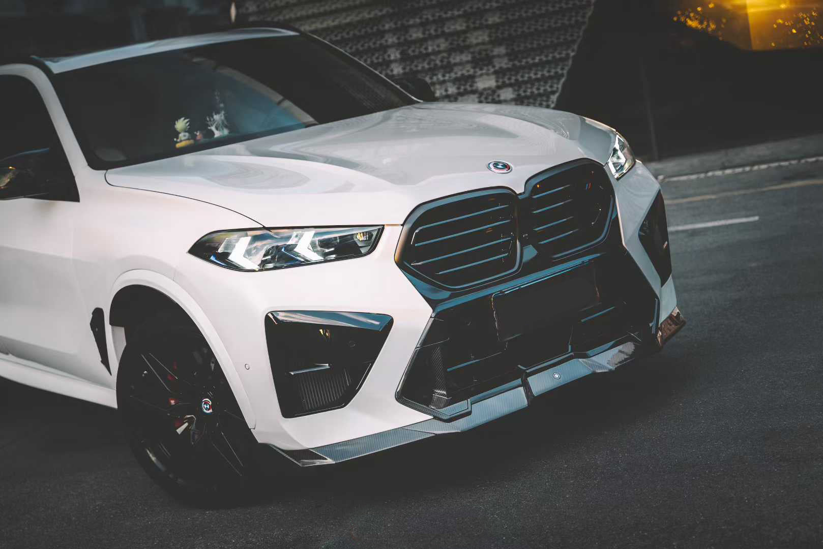 Front view of a white BMW X5M featuring a REVOZPORT carbon fiber front lip and aggressive front bumper aero styling parked on an urban street at night.