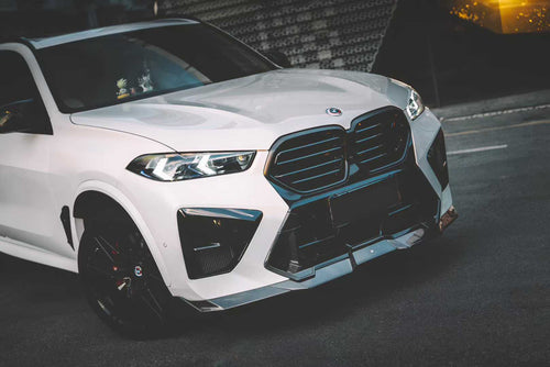 Front view of a white BMW X5M featuring a REVOZPORT carbon fiber front lip and aggressive front bumper aero styling parked on an urban street at night.