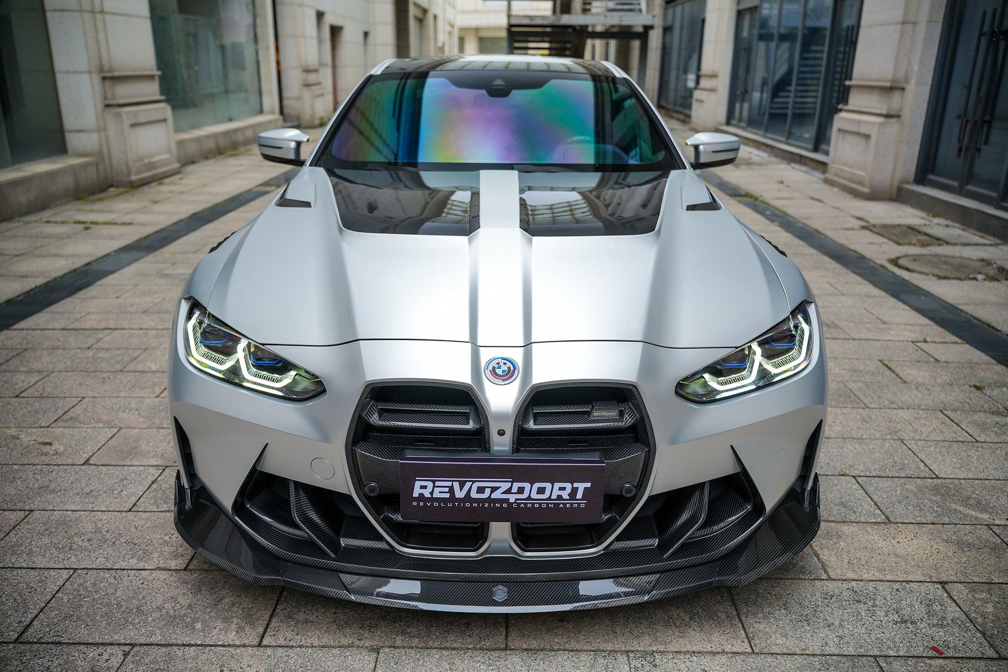 Silver BMW G82 M4 featuring a REVOZPORT carbon fiber body kit with front lip, side skirts, vented fenders, and aggressive aero styling parked in a modern urban courtyard.