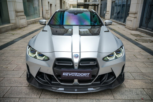Silver BMW G82 M4 featuring a REVOZPORT carbon fiber body kit with front lip, side skirts, vented fenders, and aggressive aero styling parked in a modern urban courtyard.