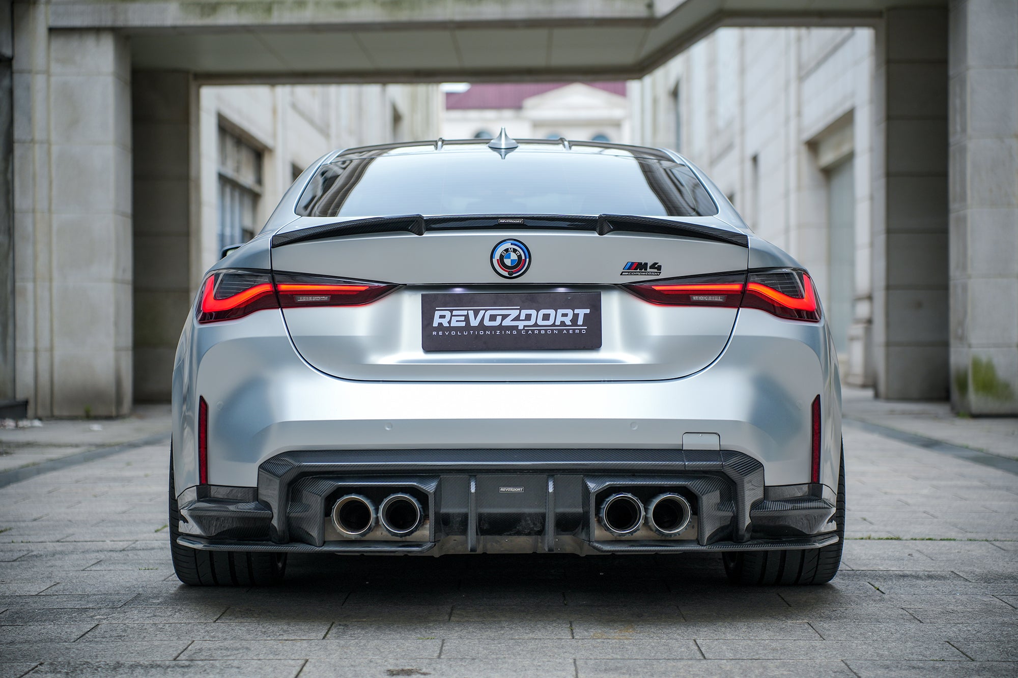 Silver BMW G82 M4 featuring a REVOZPORT carbon fiber body kit with front lip, side skirts, vented fenders, and aggressive aero styling parked in a modern urban courtyard.