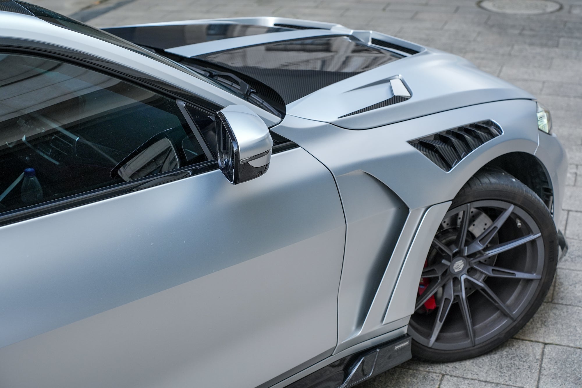 Silver BMW G82 M4 featuring a REVOZPORT carbon fiber body kit with front lip, side skirts, vented fenders, and aggressive aero styling parked in a modern urban courtyard.