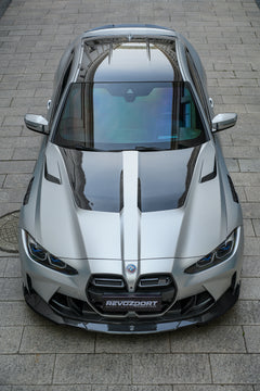 Silver BMW G82 M4 featuring a REVOZPORT carbon fiber body kit with front lip, side skirts, vented fenders, and aggressive aero styling parked in a modern urban courtyard.