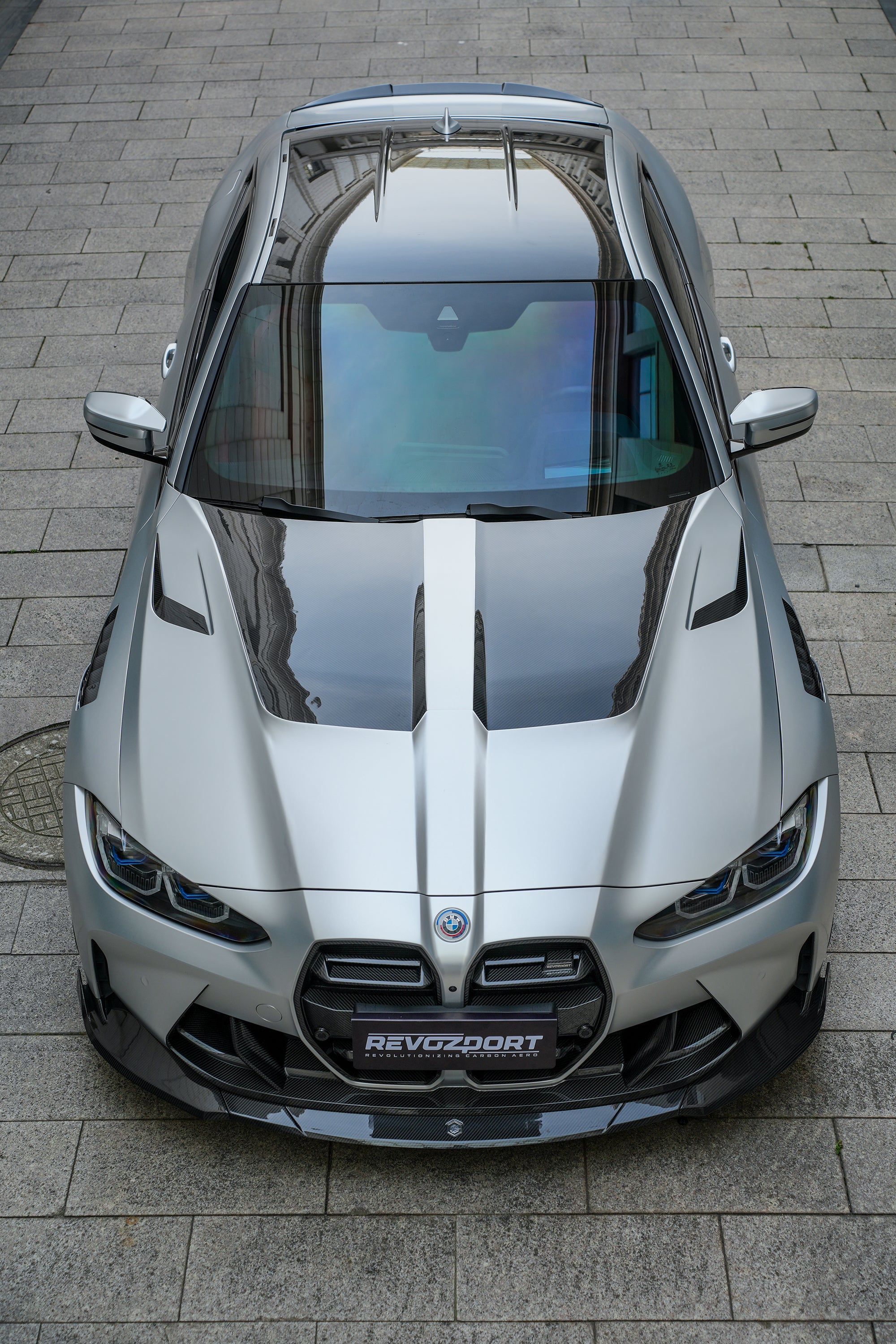 Silver BMW G82 M4 featuring a REVOZPORT carbon fiber body kit with front lip, side skirts, vented fenders, and aggressive aero styling parked in a modern urban courtyard.