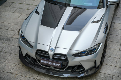 Silver BMW G82 M4 featuring a REVOZPORT carbon fiber body kit with front lip, side skirts, vented fenders, and aggressive aero styling parked in a modern urban courtyard.