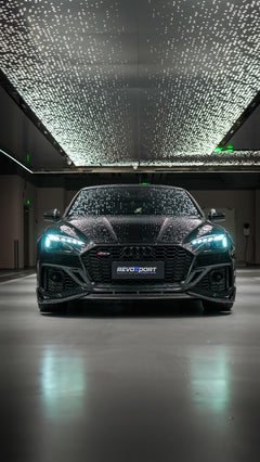 Front view of a black Audi RS5 Sportback featuring a REVOZPORT carbon fiber body kit with aggressive front lip, side aero details, and illuminated LED headlights inside a modern luxury garage.
