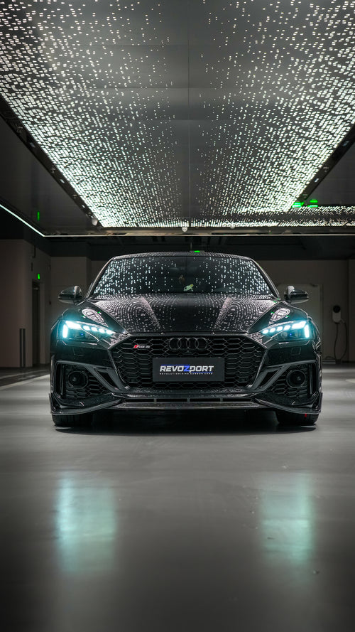 Front view of a black Audi RS5 Sportback featuring a REVOZPORT carbon fiber body kit with aggressive front lip, side aero details, and illuminated LED headlights inside a modern luxury garage.