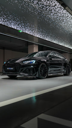 Front view of a black Audi RS5 Sportback featuring a REVOZPORT carbon fiber body kit with aggressive front lip, side aero details, and illuminated LED headlights inside a modern luxury garage.