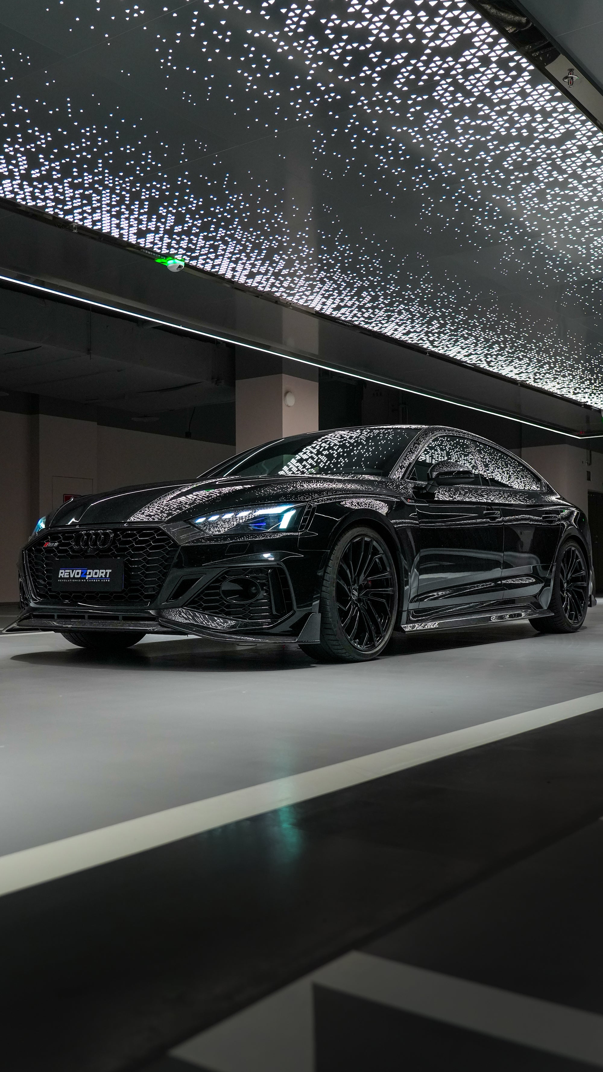 Front view of a black Audi RS5 Sportback featuring a REVOZPORT carbon fiber body kit with aggressive front lip, side aero details, and illuminated LED headlights inside a modern luxury garage.