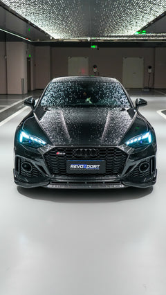 Front view of a black Audi RS5 Sportback featuring a REVOZPORT carbon fiber body kit with aggressive front lip, side aero details, and illuminated LED headlights inside a modern luxury garage.
