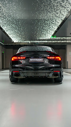 Front view of a black Audi RS5 Sportback featuring a REVOZPORT carbon fiber body kit with aggressive front lip, side aero details, and illuminated LED headlights inside a modern luxury garage.