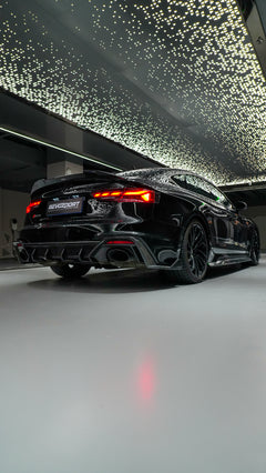 Front view of a black Audi RS5 Sportback featuring a REVOZPORT carbon fiber body kit with aggressive front lip, side aero details, and illuminated LED headlights inside a modern luxury garage.
