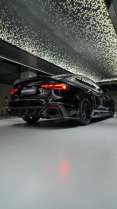 Front view of a black Audi RS5 Sportback featuring a REVOZPORT carbon fiber body kit with aggressive front lip, side aero details, and illuminated LED headlights inside a modern luxury garage.