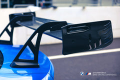 Blue BMW G80 M3 with REVOZPORT carbon fiber front splitter, side skirts, vented fenders, and large rear wing parked in a landscaped outdoor plaza.