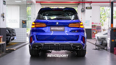 Blue BMW X5M featuring an aggressive REVOZPORT carbon fiber body kit with front lip, side skirts, enlarged front air intakes, and sporty aero styling displayed inside a modern performance workshop.