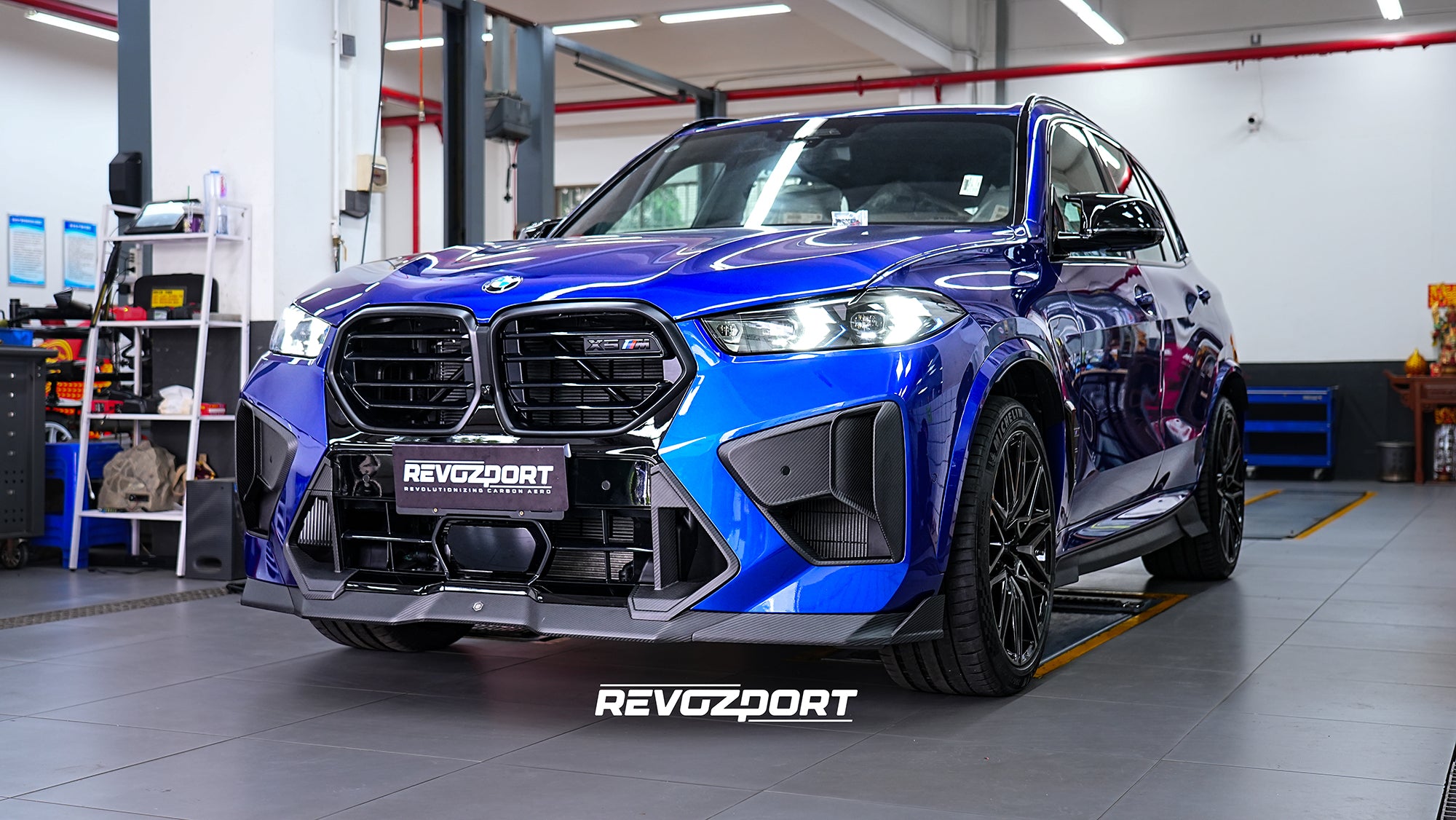 Blue BMW X5M featuring an aggressive REVOZPORT carbon fiber body kit with front lip, side skirts, enlarged front air intakes, and sporty aero styling displayed inside a modern performance workshop.