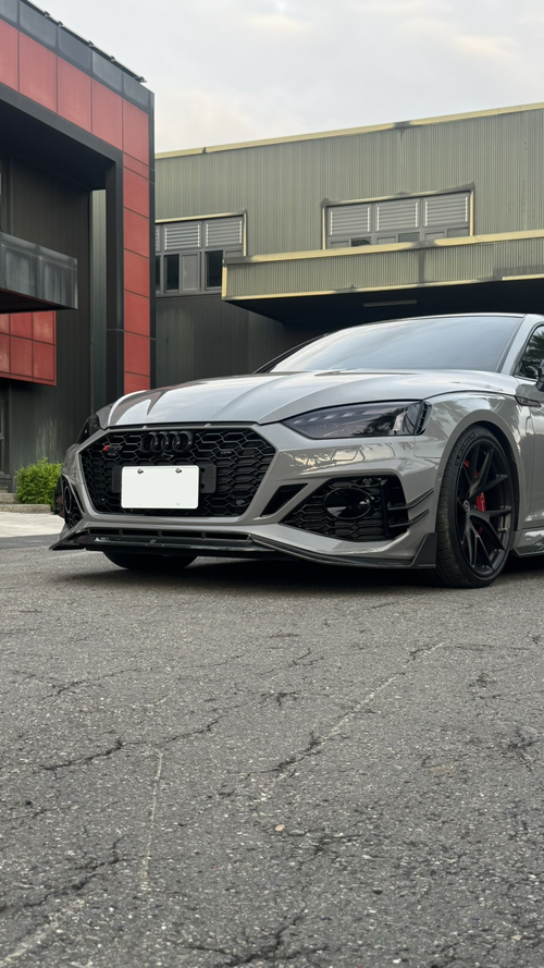 Gray Audi RS5 Sportback featuring a REVOZPORT carbon fiber front lip, side skirts, and aggressive front aero styling parked outdoors near tropical landscaping.