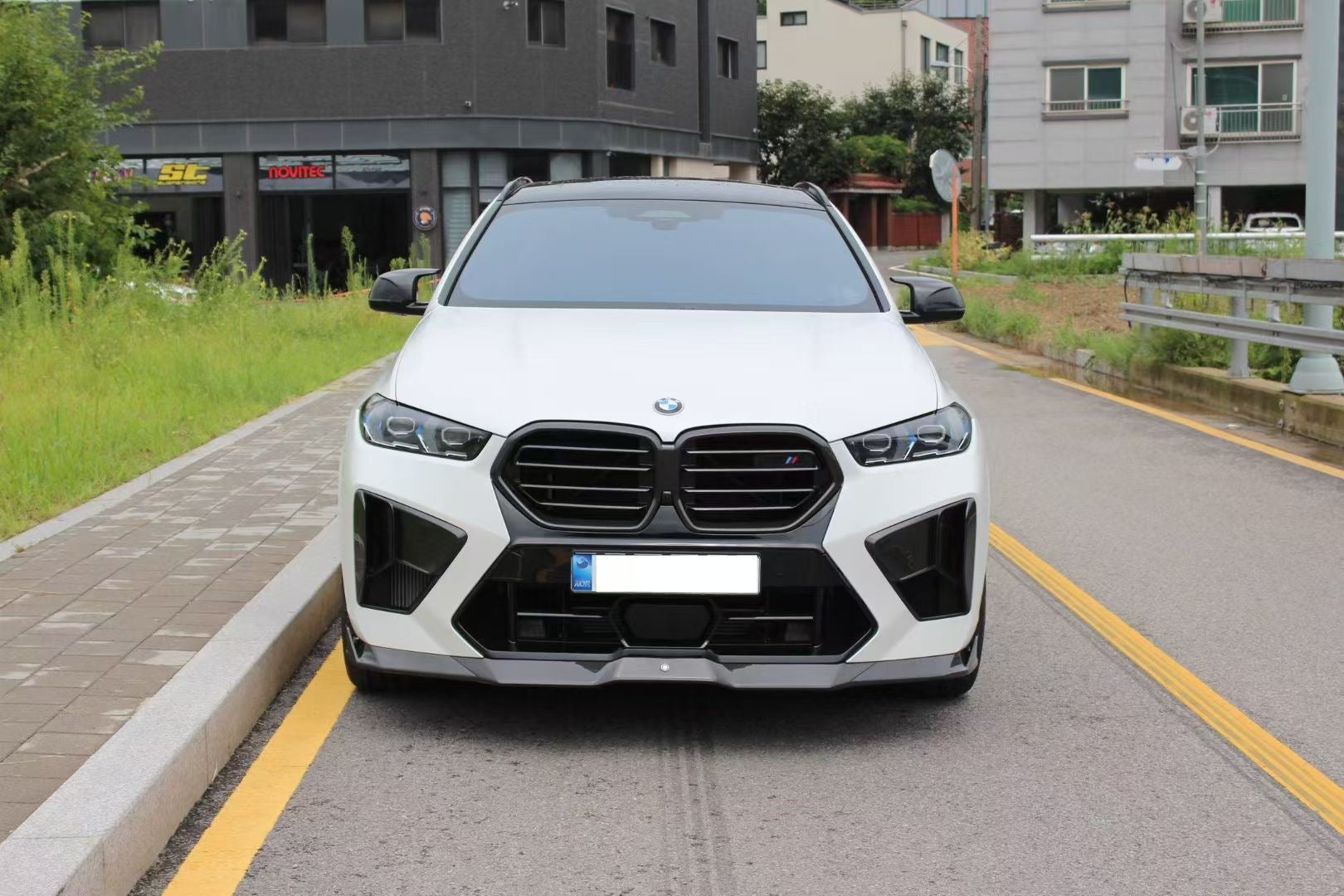 Front view of a white BMW X6M featuring an aggressive REVOZPORT front bumper and carbon fiber aero styling parked on a quiet urban street.