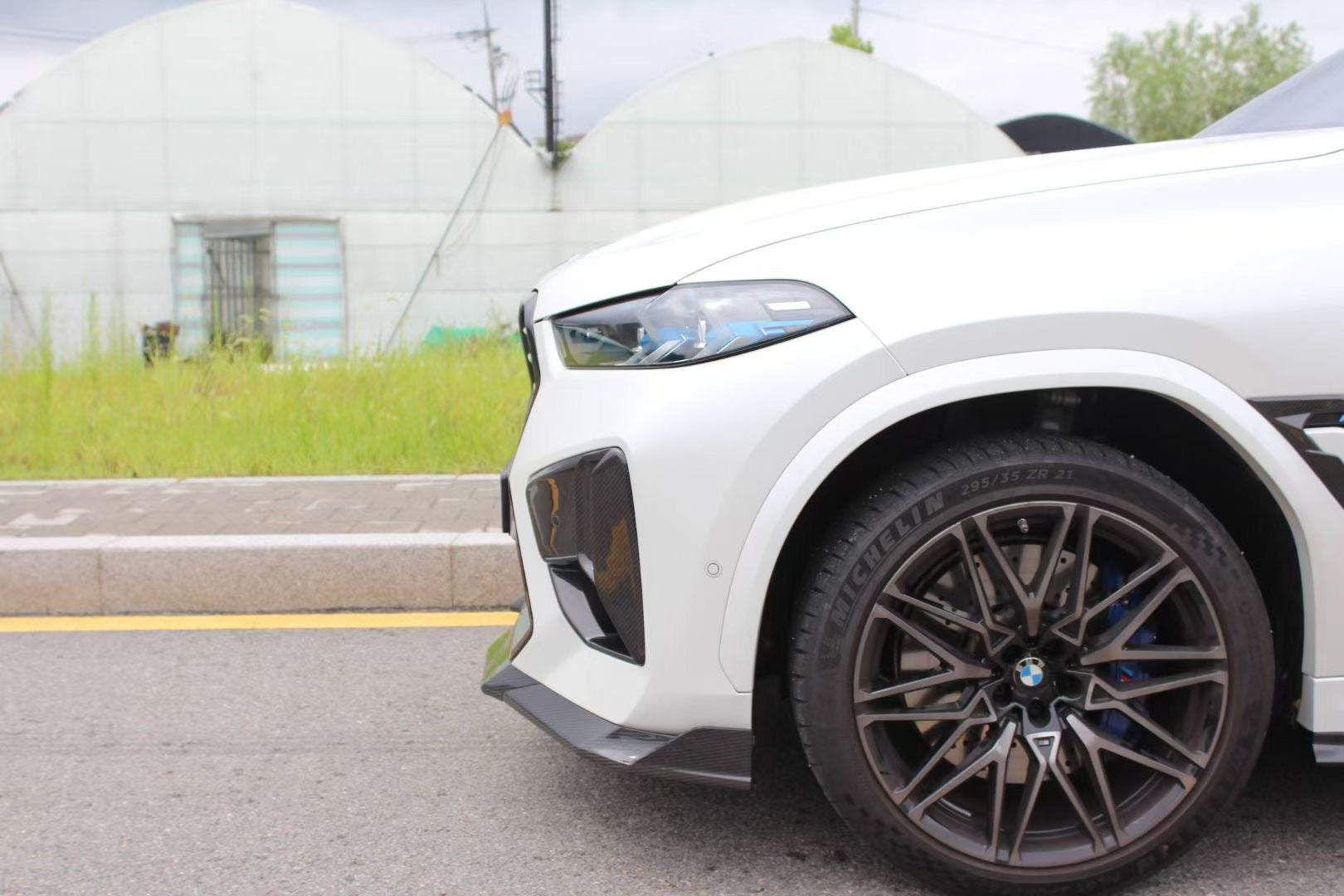 Front view of a white BMW X6M featuring an aggressive REVOZPORT front bumper and carbon fiber aero styling parked on a quiet urban street.