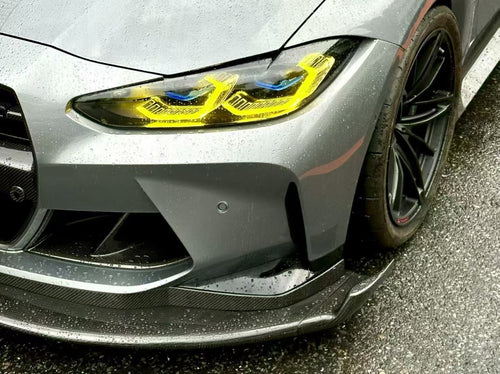 Gray BMW G80 M3 with aggressive carbon fiber aero kit, front splitter, side skirts, and rear wing parked on a rainy urban street with yellow DRL headlights illuminated.