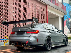 Gray BMW G80 M3 with aggressive carbon fiber aero kit, front splitter, side skirts, and rear wing parked on a rainy urban street with yellow DRL headlights illuminated.