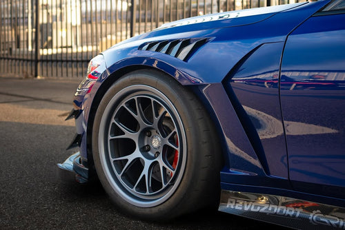 Close-up of a blue wide-body performance car featuring aggressive aerodynamic fender vents, extended side skirts, and lightweight multi-spoke wheels. The image highlights the custom bodywork, low-profile performance tire, and carbon fiber aero details in an outdoor automotive setting.