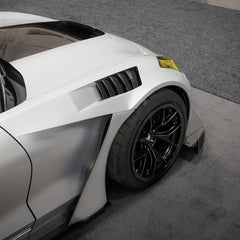 Close-up of a silver performance car’s front wheel and brake setup, featuring black forged multi-spoke wheels, large drilled brake rotors, and high-performance racing tires. The image also highlights the aggressive wide-body fender design and carbon fiber aerodynamic components in a motorsport environment.