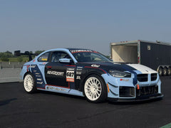 A light blue and white BMW race car parked in a racetrack paddock, featuring a wide-body aerodynamic kit, carbon fiber front splitter, large air intakes, and white racing wheels. The car is covered with sponsor decals and track graphics, with racetrack barriers and trees visible in the background.