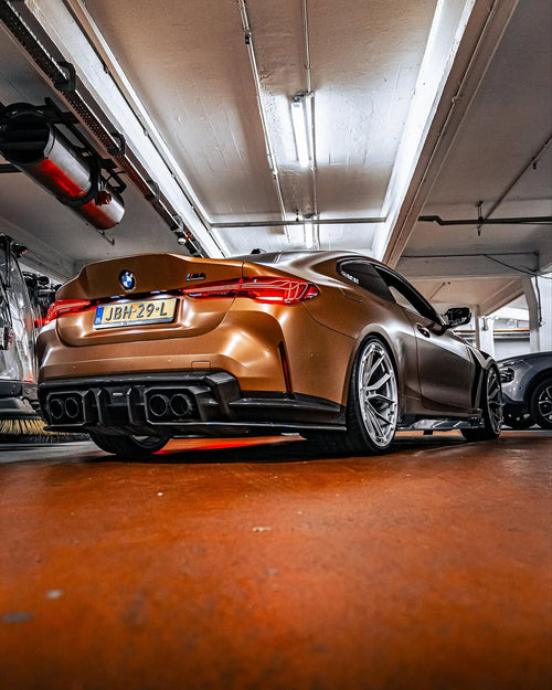 Bronze BMW G82 M4 fitted with REVOZPORT forged silver multi-spoke wheels, parked on a luxury city street alongside a matching Audi RS model.