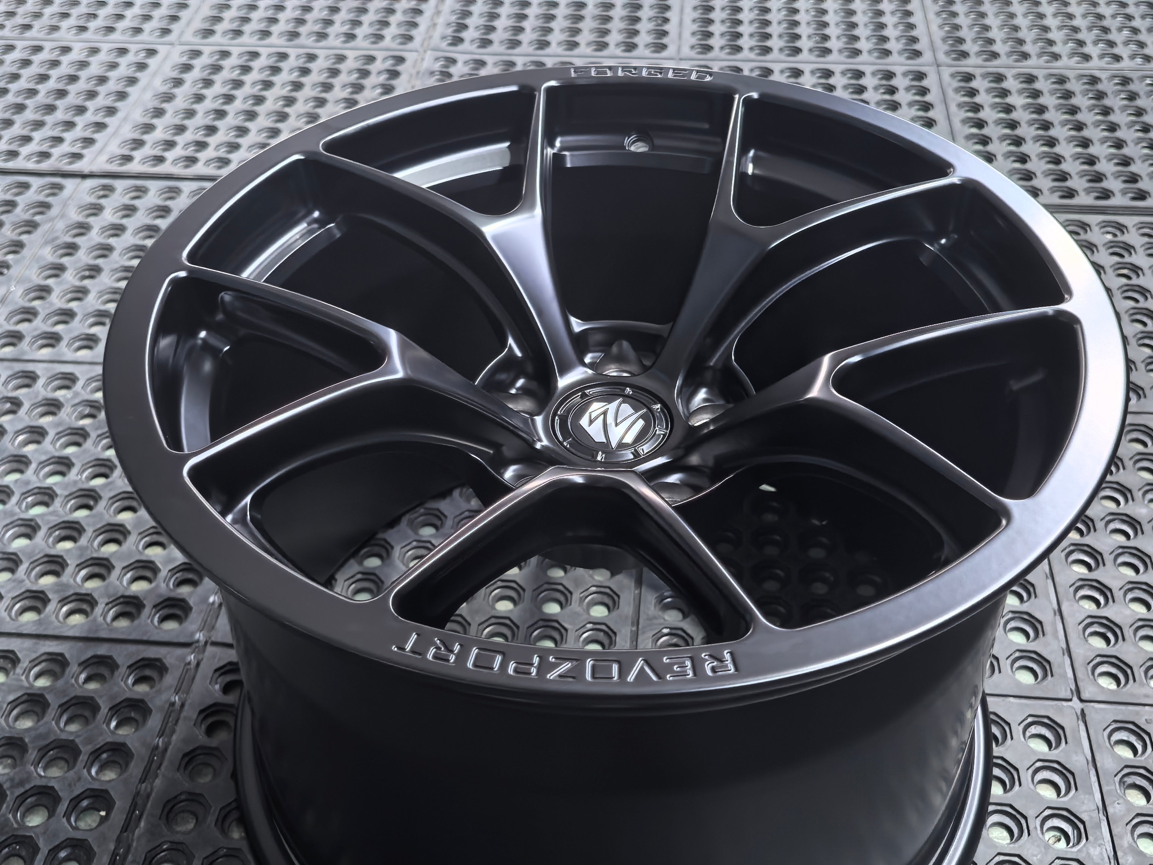 Studio-style close-up of a matte black forged performance wheel featuring a lightweight multi-spoke design and REVOZPORT branding. The wheel is displayed on a textured industrial floor, highlighting its premium finish, deep concave profile, and motorsport-inspired styling.