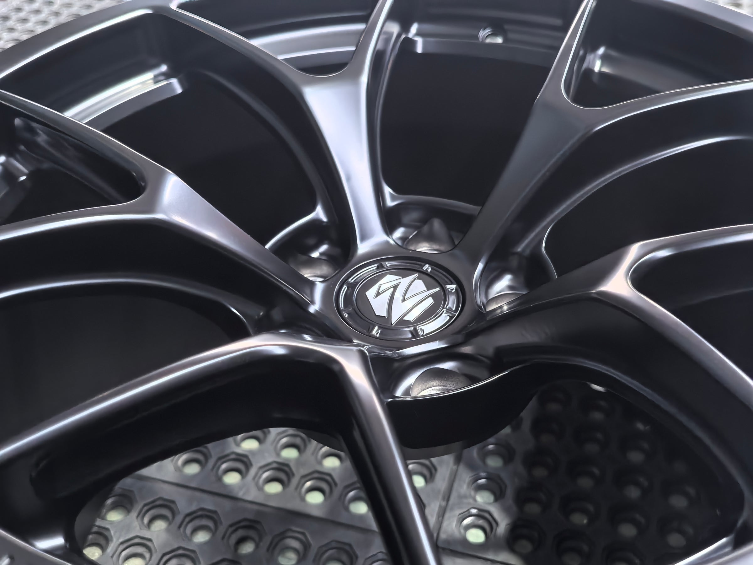 Studio-style close-up of a matte black forged performance wheel featuring a lightweight multi-spoke design and REVOZPORT branding. The wheel is displayed on a textured industrial floor, highlighting its premium finish, deep concave profile, and motorsport-inspired styling.